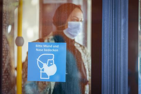 Eine Frau mit einer Mund-Nasen-Bedeckung steht in einem Bus. Ein Schild weist darauf hin, dass im ÖPNV Maskenpflicht gilt. Foto: Sascha Kopp