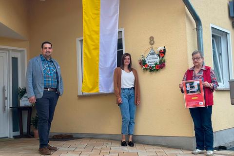 Eine besondere Aktion zum 725-jährigen Bestehen von Waldernbach: Karsten Gerhard (v. l.), Carolin Reitz und Renate Schuld zeigen den Fahnenschmuck am Haus sowie das gestaltete Ortsnamenschild. Jeder Waldernbacher ist an Fronleichnam dazu aufgerufen, sein Haus ebenfalls zu schmücken.  Foto: Ortsbeirat Waldernbach 